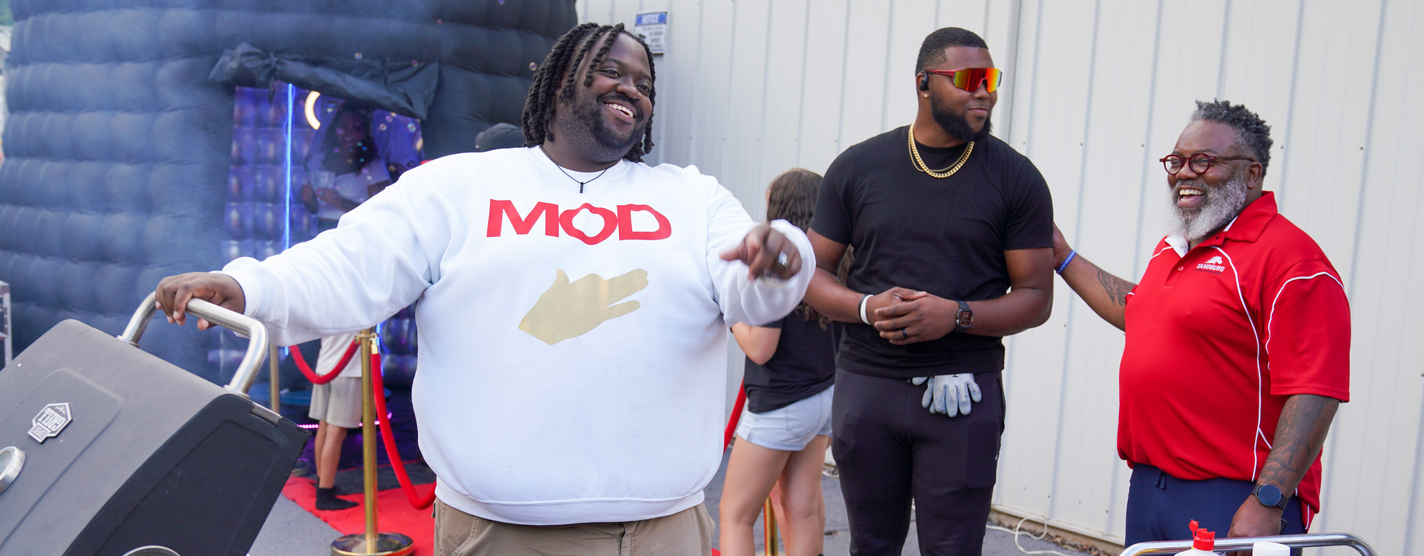 Three men standing talking around a grill, one has a MOD shirt on.