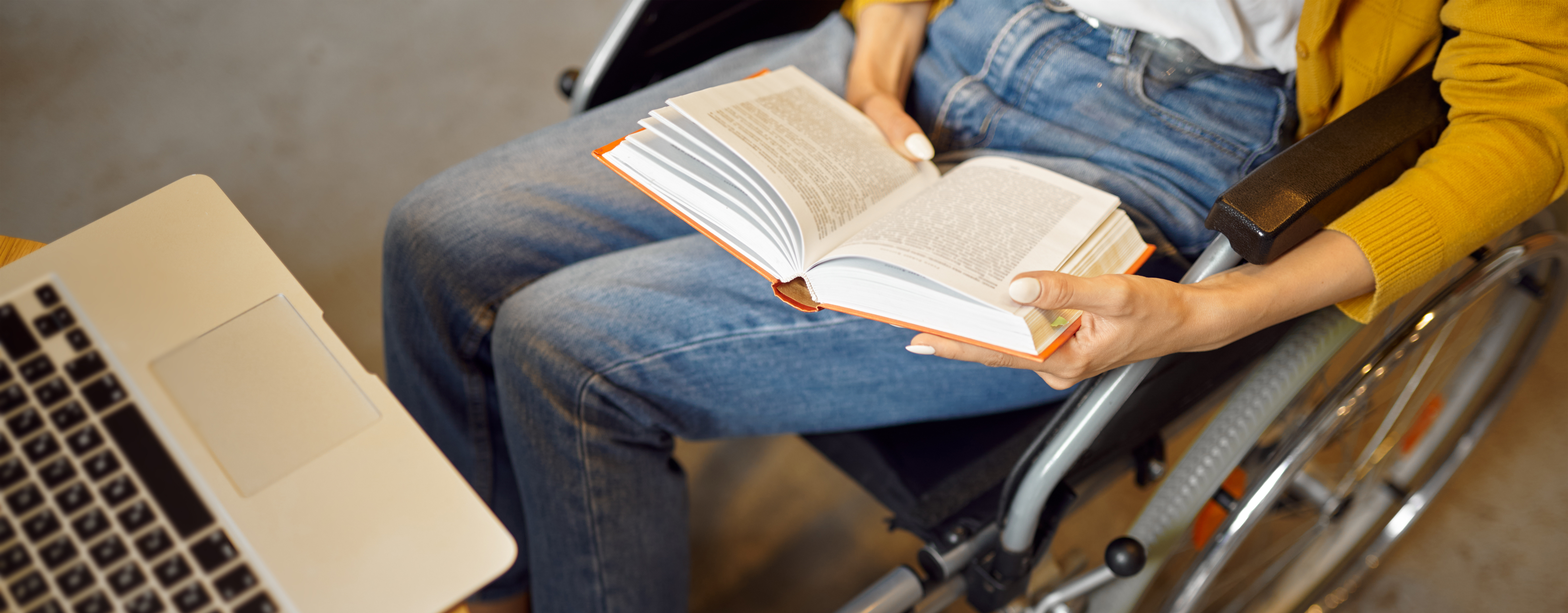 student in wheelchair reading a book with a laptop on the table in front of her.