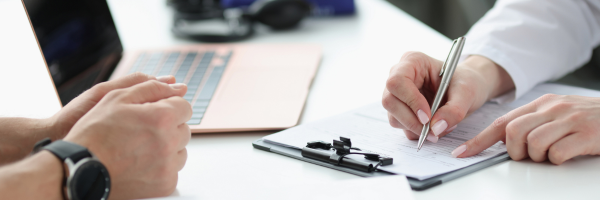 two people siting at a desk one of them has a pen and page filing a form out.